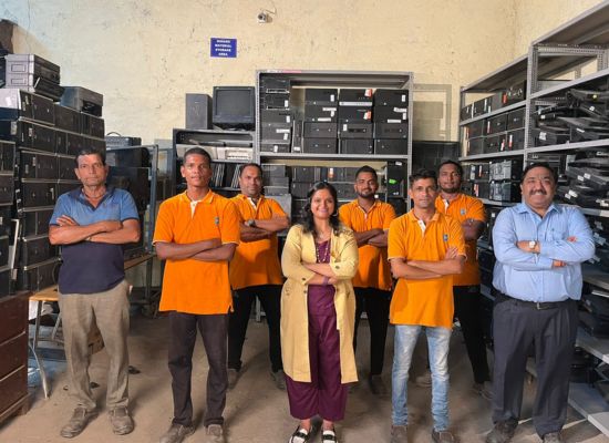 Indian people standing in an archive, facing the camera, smiling. A woman in the middle, surrounded by five men in orange T-Shirts (next to and behind her) as well as two other men in blue shirts to each side of the group.