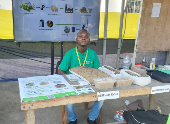 An african man sitting behind a table with samples of his products, smiling into camera